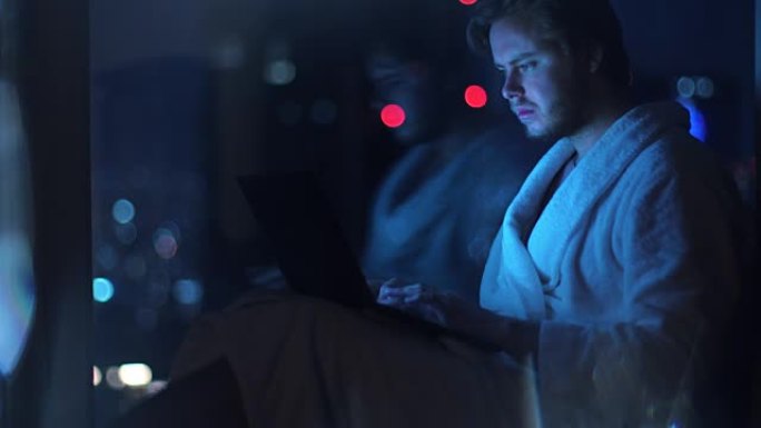 穿着浴袍的旅行年轻企业家使用笔记本电脑，完成工作并在窗户旁放松。聪明的年轻商人在大城市，放松。