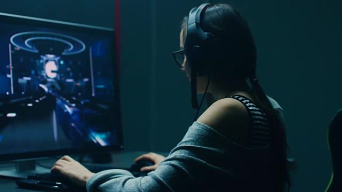 在美丽的职业玩家女孩坐下来在她的个人电脑上玩第一人称射击在线视频游戏的镜头之后。休闲可爱的怪胎戴着眼