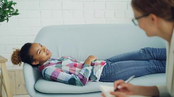 细心的心理学家正在与非裔美国妇女进行咨询，在客户躺在沙发上微笑着说话时聆听并做笔记。