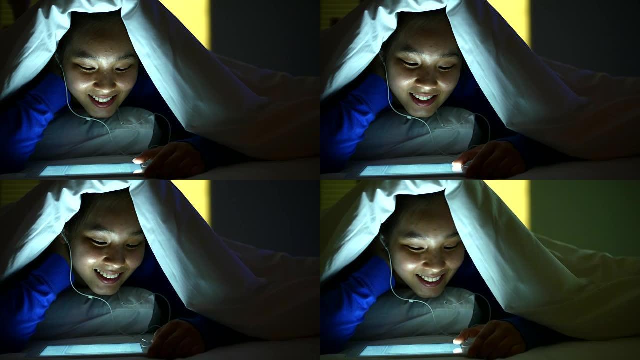 亚洲年轻女子晚上在床罩下的数字平板电脑上听音乐，4k(UHD)