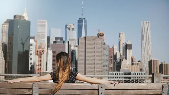 快乐的女性旅行者的后景，长发在风中飘扬，在长凳上欣赏令人惊叹的曼哈顿天际线景色