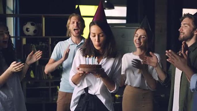 快乐的年轻女士拿着生日蛋糕和吹蜡烛的慢动作站在办公室，同事们鼓掌祝贺她。