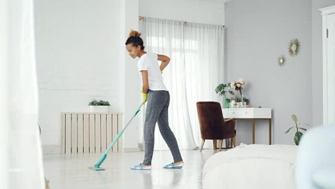 年轻的非洲裔美国妇女正在用拖把打扫现代房屋的地板，漂亮的女孩穿着家居服，拖鞋和橡胶手套专心工作。
