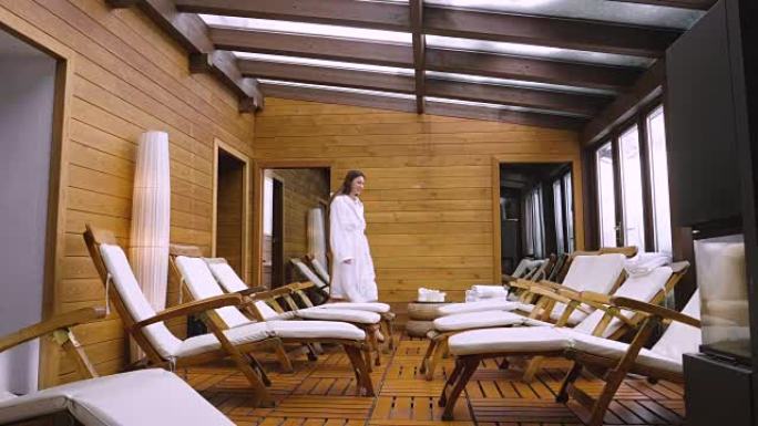 一个美丽的女人带着白色浴袍进入木制水疗中心 (健康中心) 的休闲室，坐在其中一个躺椅上放松。