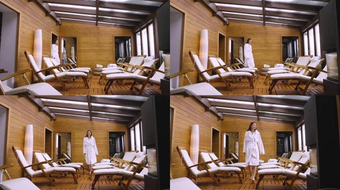一个美丽的女人带着白色浴袍进入木制水疗中心 (健康中心) 的休闲室，坐在其中一个躺椅上放松。
