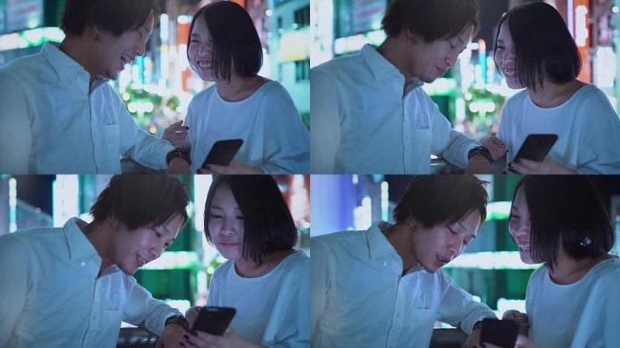 年轻的东日本男孩和女孩夫妇聊天，大笑，使用手机和共享屏幕。在背景中模糊的广告广告牌和夜晚的城市灯光。