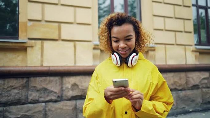 快乐的非洲裔美国学生正在使用智能手机浏览或发短信的朋友站在现代城市的街道上，戴着耳机微笑。