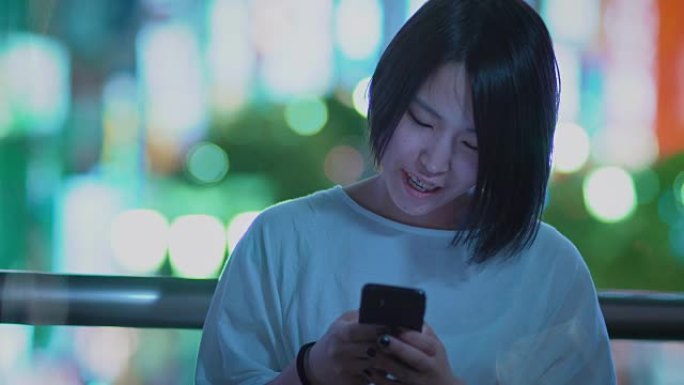 穿着休闲服的迷人日本女孩的肖像使用智能手机。在后台，大城市的广告广告牌灯光在夜晚发光。