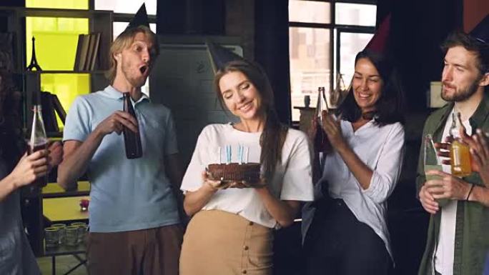 公司员工正在庆祝生日，女人拿着蛋糕吹蜡烛，她的同事在拍手，用饮料碰杯。