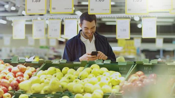 在超市: 男人站在新鲜农产品区附近选择水果时使用智能手机。大光明正宗商店。