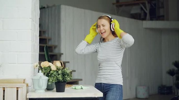 好看的女孩正在家里给桌子除尘，通过耳机听音乐，开心地唱歌跳舞。千禧一代，家庭和室内概念。