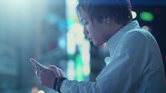 使用智能手机的英俊另类日本男孩的肖像。在后台，大城市的广告广告牌灯光在夜晚发光。