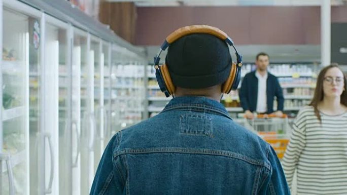 在超市:时尚的非洲裔美国人戴着耳机走过商店的冷冻商品区。后面的视图拍摄。缓慢的运动。