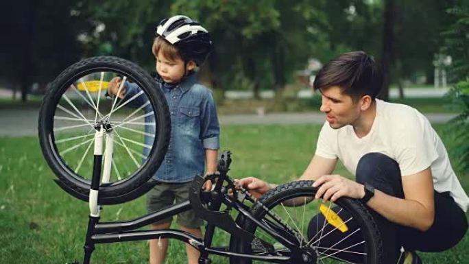 夏天，当父亲在公园的草坪上与他交谈时，戴头盔的好奇孩子正在旋转自行车车轮和踏板。家庭、休闲和积极的生