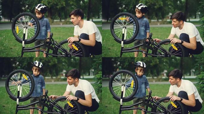 夏天，当父亲在公园的草坪上与他交谈时，戴头盔的好奇孩子正在旋转自行车车轮和踏板。家庭、休闲和积极的生