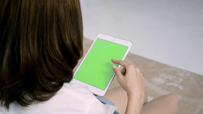 年轻的亚洲妇女使用绿色屏幕的黑色平板电脑设备。亚洲妇女拿着平板电脑，坐在客厅的沙发上滚动页面。色度键