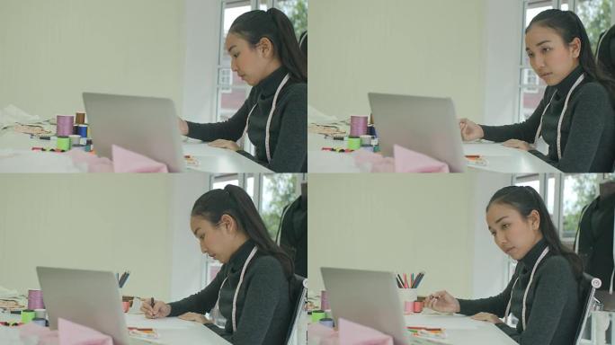 DS MS美丽的亚洲女性时装设计师坐在办公桌前看着现代工作室笔记本电脑上的草图，做笔记