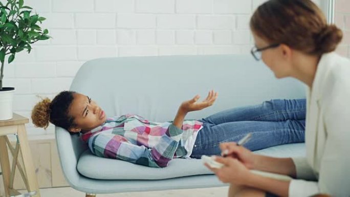 沮丧的非裔美国女孩正在与躺在沙发上的心理学家交谈，而戴着眼镜的治疗师正在听和写。帮助和专家概念。