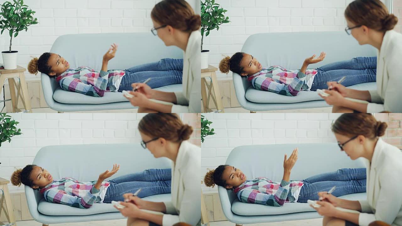 沮丧的非裔美国女孩正在与躺在沙发上的心理学家交谈，而戴着眼镜的治疗师正在听和写。帮助和专家概念。