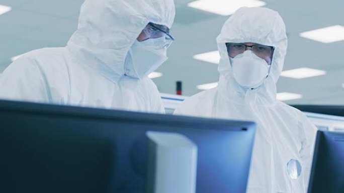 两名穿着无菌服的科学家/技术人员在个人计算机上检查数据，进行了讨论。他们在药物制造实验室工作。