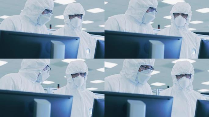 两名穿着无菌服的科学家/技术人员在个人计算机上检查数据，进行了讨论。他们在药物制造实验室工作。