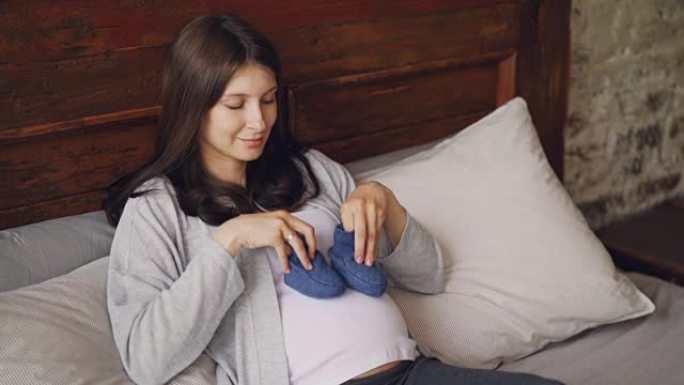 顽皮的期待母亲在怀孕的肚子上穿着蓝色婴儿鞋，在阁楼风格的卧室里躺在家里的床上微笑。怀孕和孕产概念。
