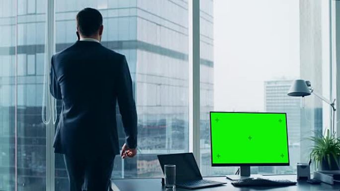 自信的商人在办公室打电话，桌上的个人电脑显示绿色色度键屏幕。在窗户上全景大城市的景色。