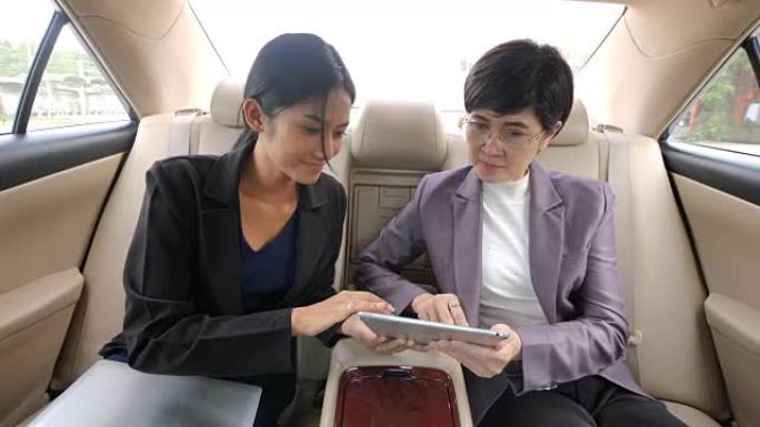 两个女商人坐在汽车后座上，用数字平板电脑聊天