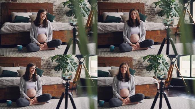美丽的年轻女子博主正在录制有关怀孕的视频，她的vlog坐在家里的地板上，在三脚架上聊天并看着智能手机