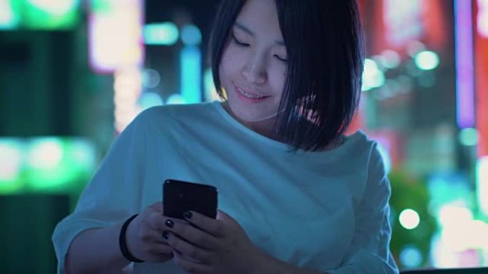 穿着休闲服的迷人日本女孩的肖像使用智能手机，在城市中漫步。在后台，大城市的广告广告牌灯光在夜晚发光。
