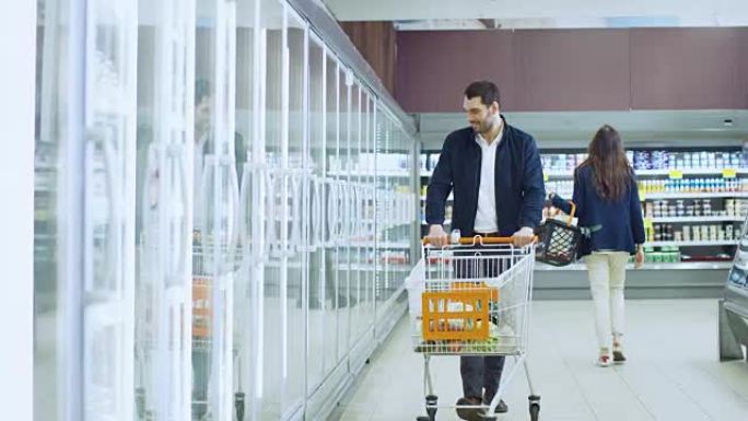 在超市: 帅哥推购物卡，在冷冻商品区浏览商品。男人看着玻璃门冰箱，选择乳制品。其他顾客购物。
