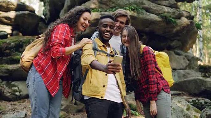 快乐的非裔美国人家伙正在与朋友一起在森林徒步旅行期间使用智能手机进行视频通话，年轻人正在看着屏幕，挥