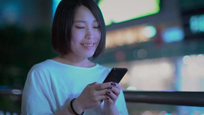 穿着休闲服的迷人另类日本女孩的肖像使用智能手机，在城市中漫步。在后台，大城市的广告广告牌灯光在夜晚发