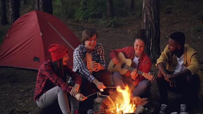 漂亮女人正在弹吉他，而她的朋友们正在唱着有趣的歌曲，笑着煮着棉花糖着火，拍手。音乐、自然和有趣的概念