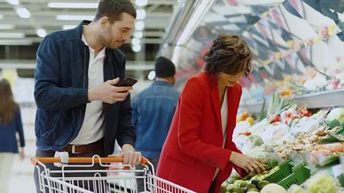 在超市: 幸福的夫妇购物，在新鲜农产品区选择水果和蔬菜。男人在智能手机上指向清单并推动购物车，女人选