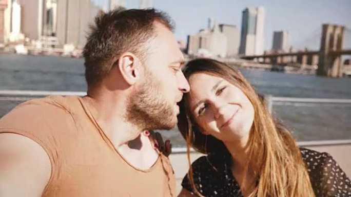 微笑着快乐的高加索年轻夫妇站在史诗般的纽约天际线曼哈顿景色，亲吻并自拍