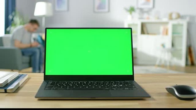 现代笔记本电脑，绿色模拟屏幕显示，站在舒适的客厅的桌子上。拿着手机的男人坐在椅子上休息。