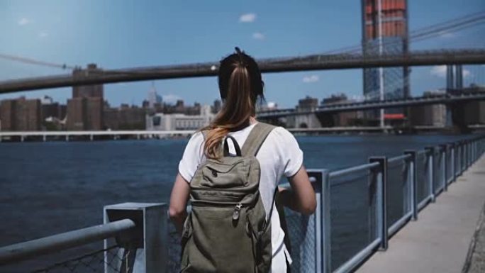 摄像机跟随年轻的女游客背着背包，享受着轻松的走向阳光河岸的布鲁克林大桥