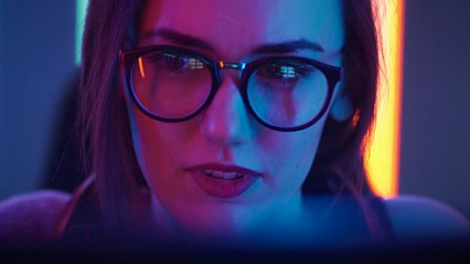 美丽的年轻女孩坐在电脑前的肖像，在互联网上浏览，玩在线游戏，流媒体。可爱的女孩戴着眼镜在凉爽的复古霓