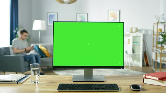 现代个人电脑，绿色模拟屏幕显示，站在舒适的客厅的桌子上。一个拿着手机的男人在椅子上放松。