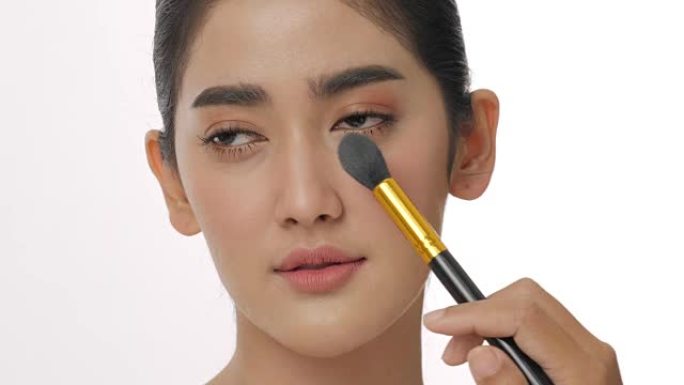 迷人的亚洲女人的特写镜头用化妆刷在脸上涂粉。专业艺术家在纯白色背景下在颧骨上涂抹遮瑕膏腮红。美丽和时