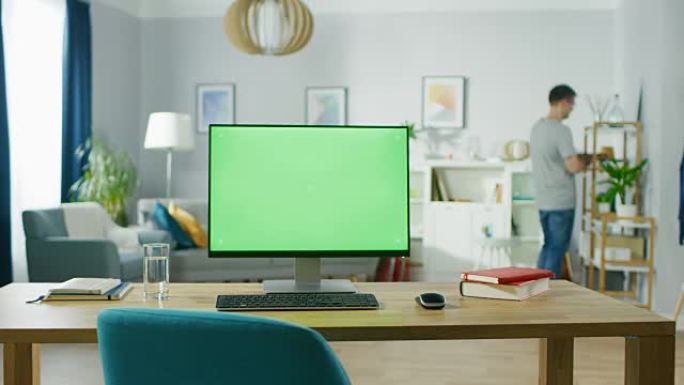 现代个人电脑，模拟绿屏显示屏站在舒适的家庭办公室的桌子上。在背景中，聪明的人拿了明亮客厅书架的书。