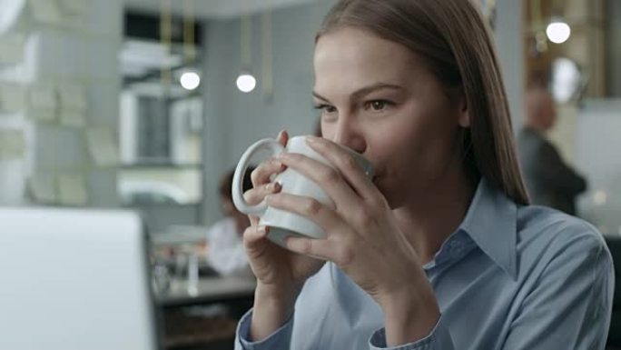年轻的女商人看着笔记本电脑喝咖啡