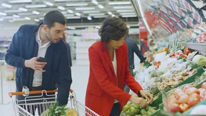 在超市: 幸福的夫妇购物，在新鲜农产品区选择水果和蔬菜。男人使用智能手机并推动购物车，女人将产品放入