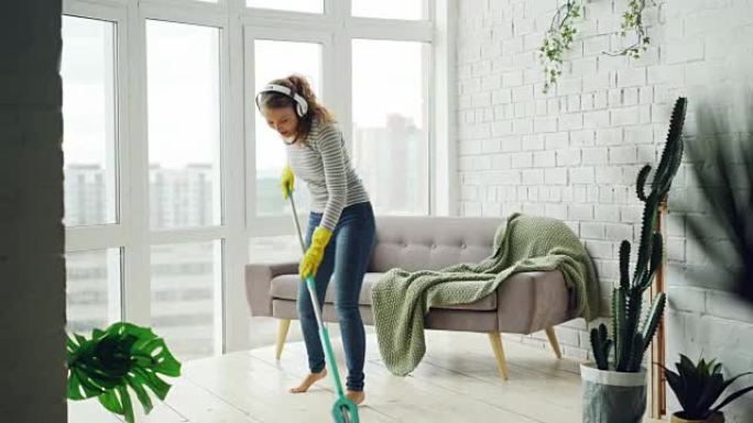 快乐的女孩在轻便的公寓里拖地板，通过耳机听音乐、跳舞和唱歌很开心。美丽的家具和植物是可见的。