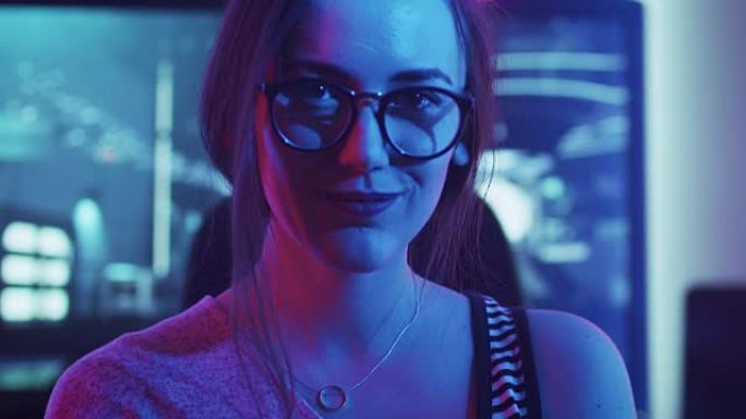 美丽的年轻职业玩家女孩坐在她的个人电脑前的肖像转过身，微笑着看着相机。迷人的极客女玩家戴着眼镜在霓虹