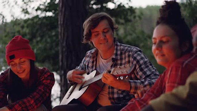 英俊的年轻人游客正在弹吉他，微笑着，而他的朋友们正在唱歌，在湖或河附近的火旁休息。绿树可见。