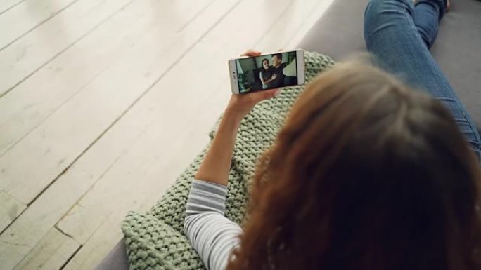 年轻的skyping女士和朋友的后视图美丽的夫妇，智能手机坐在家里的沙发上。现代技术、千禧一代和沟通