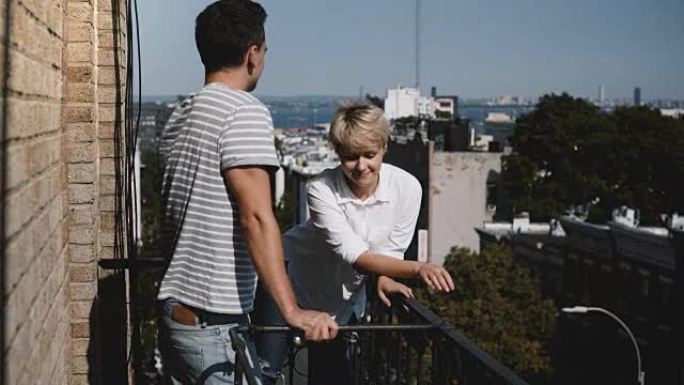 年轻的多种族浪漫夫妇站在一起，在一个阳光明媚的小阳台上聊天，可以看到令人惊叹的纽约景色