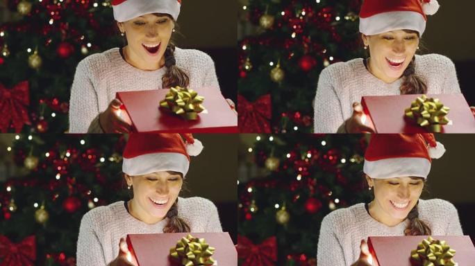 戴圣诞帽的女孩许愿并打开圣诞礼包。假期和新年的概念。这个女孩很开心，手里拿着圣诞礼物微笑着。